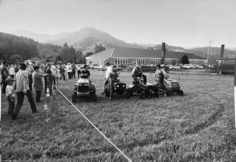 A lawnmower race on the field beside Spring Creek School, with neighbors watching along the rope line