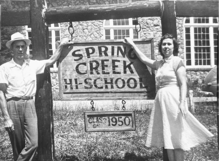 A man and a woman stand on either side of the Spring Creek Hi-School sign, with a Class of 1950 plaque below